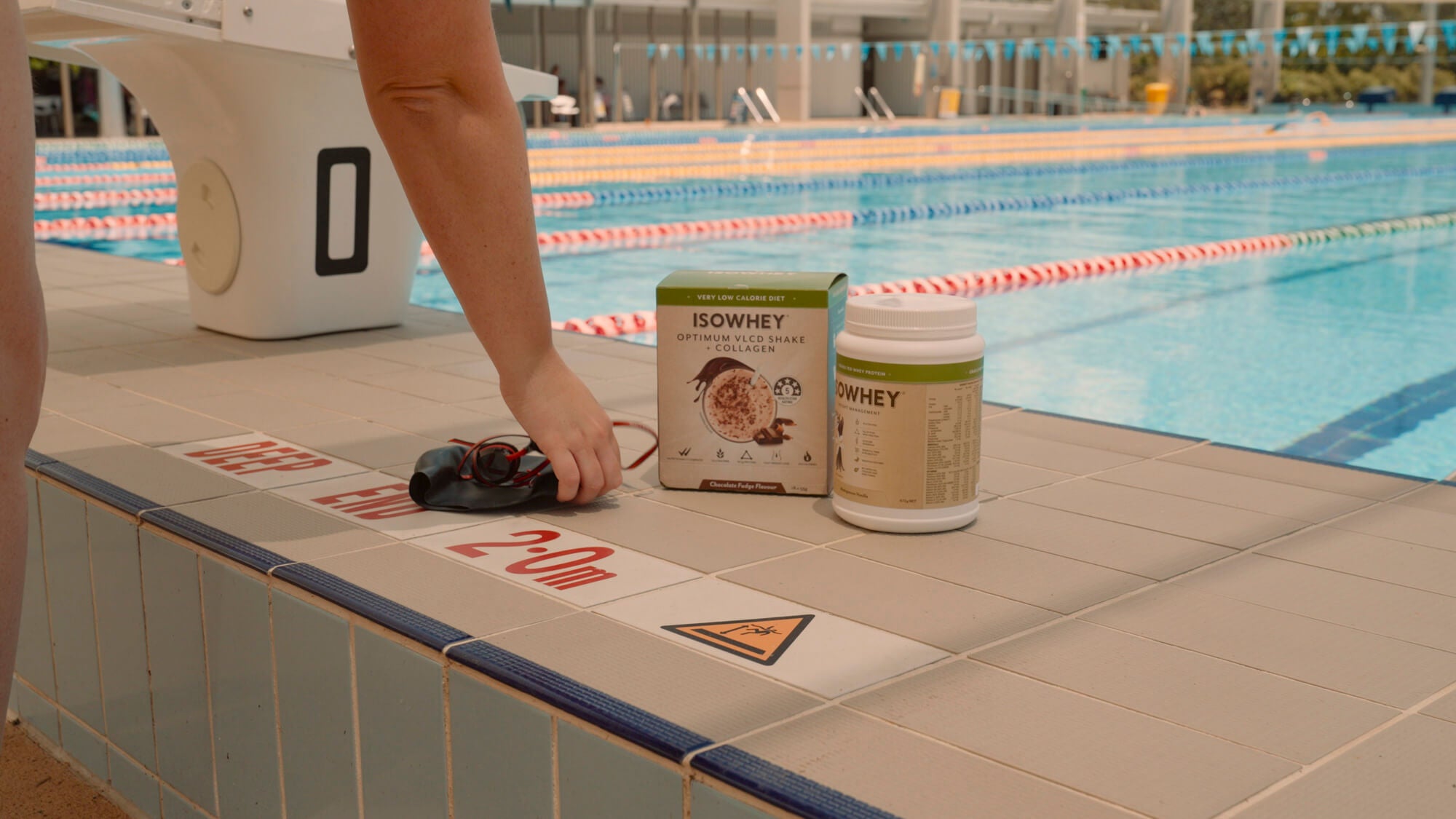 Alice reaching for her swim cap and goggles, right next to a box of IsoWhey Optimum VLCD Shake + Collagen